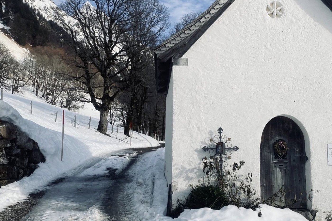 Weiße Bergkapelle mit schmiedeeisernem Wegkreuz und kahlem Bergahorn an einem Spätwintertag