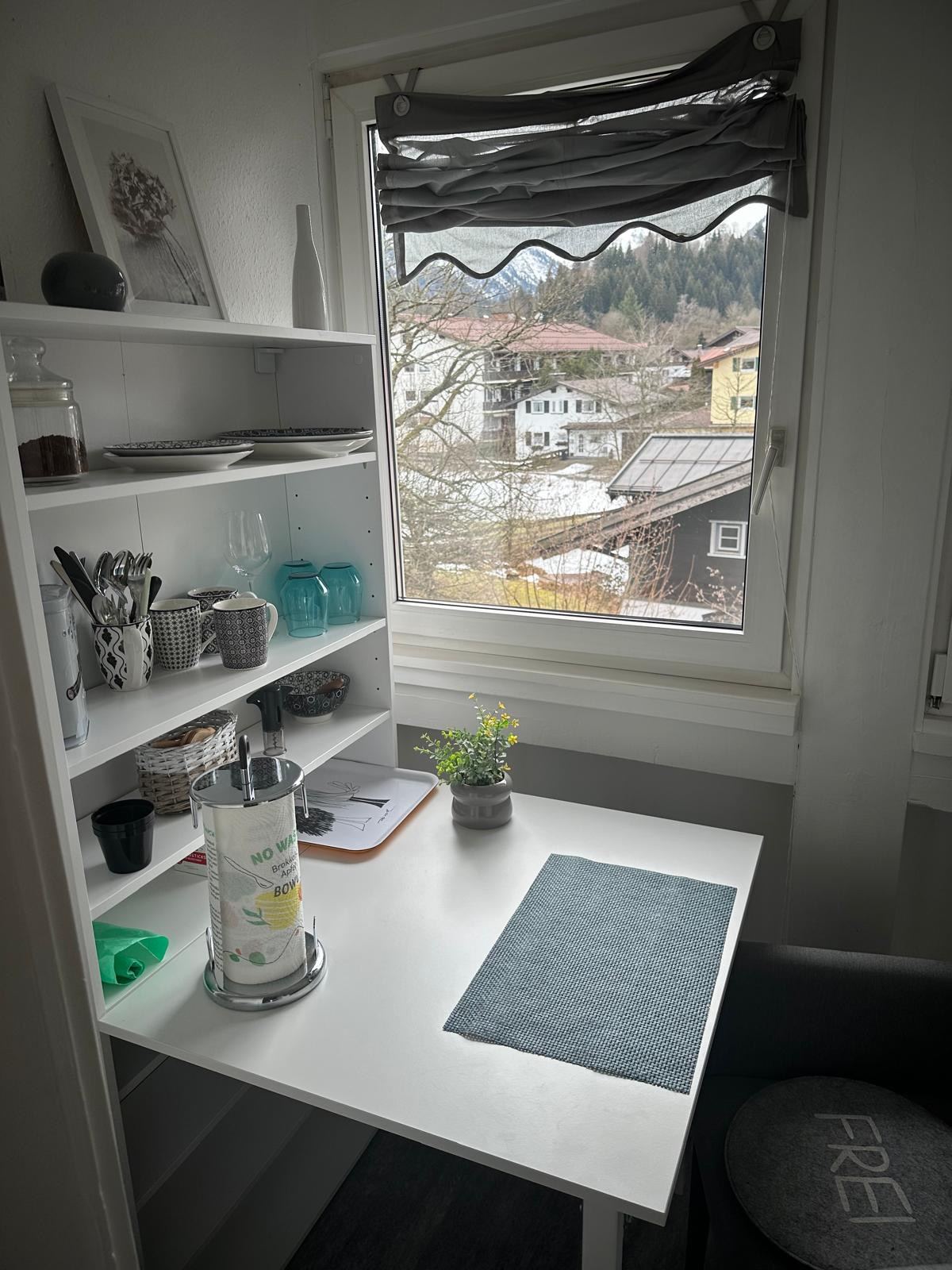Klapptisch am Fenster mit offenem Regal für Geschirr, Blick auf verschneite Dächer und Berge.