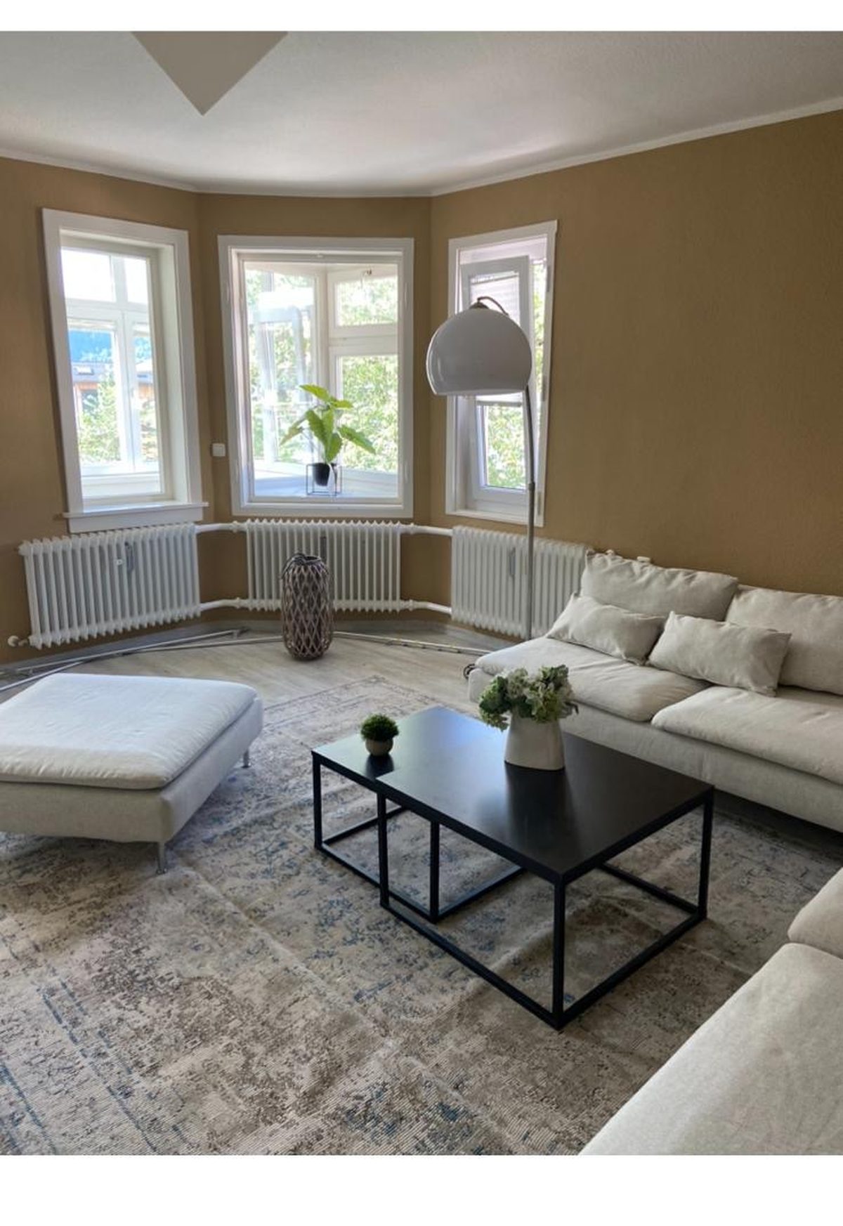 Wohnzimmer der Ferienwohnung Stillach mit Erkerfenster nach drei Seiten, heller Polstergarnitur und schwarzem Couchtisch.