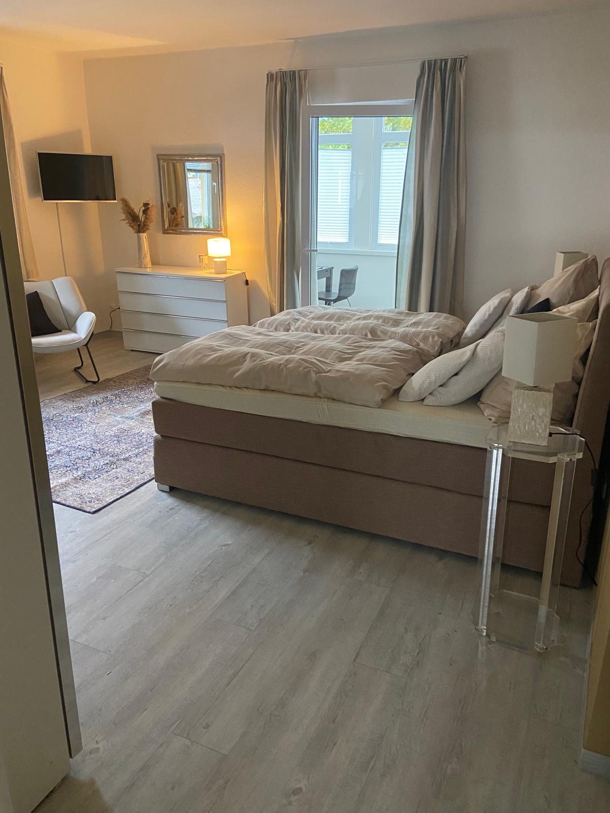 Großes Schlafzimmer mit Boxspringbett, Kommode und Sessel, Fenster zum Balkon.