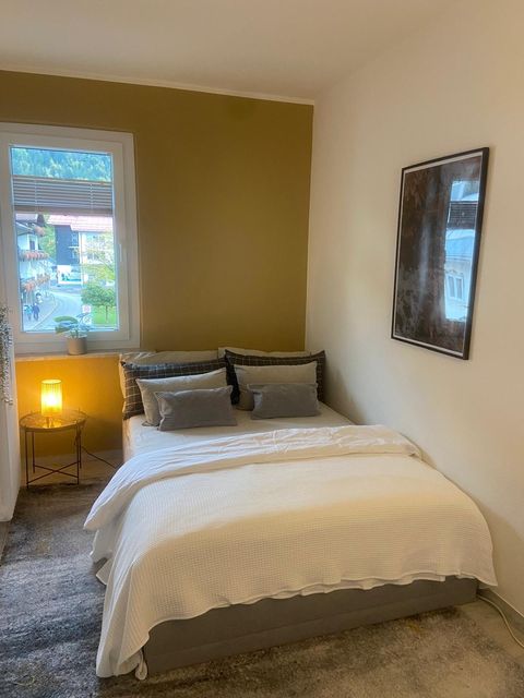 Zweites Schlafzimmer mit 140er Bett, gelber Akzentwand, Tischlampe an, Blick aus dem Fenster auf ein Allgäuer Holzhaus.